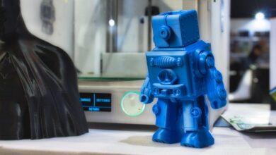 A 3D-printed blue miniature robot with intricate details and a Batman model sit on display in front of a 3D printer.