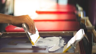 A person manually spreading opaque white ink onto a T-shirt screen stencil of the number 13 in a studio.