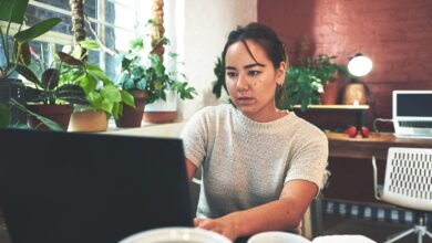 A young person sitting in their studio workspace and looking focused as they type on a large black laptop on a table.