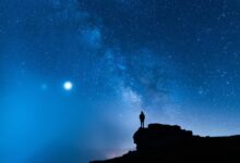 The silhouette of a single person standing on a rocky ledge in front of a deep blue sky filled with stars.