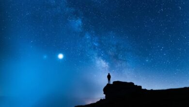 The silhouette of a single person standing on a rocky ledge in front of a deep blue sky filled with stars.
