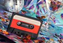 A black-and-red cassette player sitting on a surface covered in iridescent confetti alongside two disco balls and a glass.