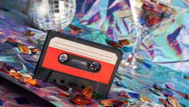 A black-and-red cassette player sitting on a surface covered in iridescent confetti alongside two disco balls and a glass.