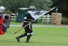 Two people, dressed as medieval knights, run across a grassy area. One holds a flag, and the other has a shield.
