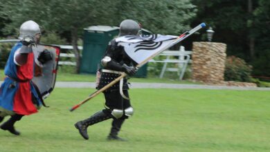 Two people, dressed as medieval knights, run across a grassy area. One holds a flag, and the other has a shield.