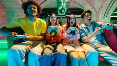A group of friends sits together on a couch, playing video games. They are in a home game room that features neon lights.