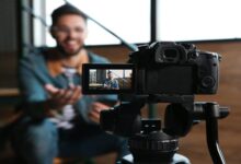 A DSLR camera on a tripod films a smiling man gesturing in an indoor setting, with the focus on the camera screen.