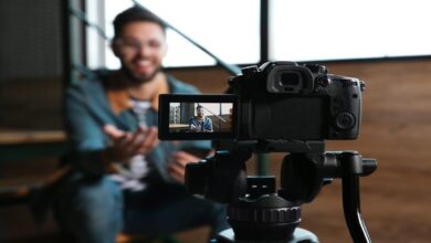 A DSLR camera on a tripod films a smiling man gesturing in an indoor setting, with the focus on the camera screen.