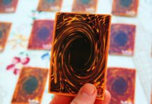 A close-up of someone holding a single Yu-Gi-Oh! card, with several others blurred in the background on a table.