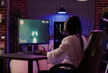 A woman sitting at her gaming setup on her desk. There is a computer tower and a game playing on the screen.