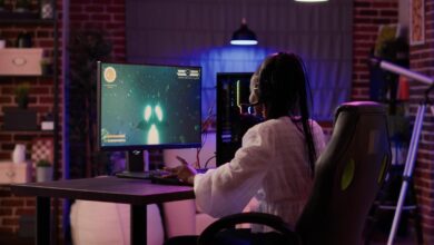 A woman sitting at her gaming setup on her desk. There is a computer tower and a game playing on the screen.