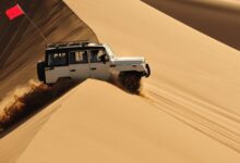 A white off-road SUV drives over steep desert sand dunes, kicking up sand, with a red safety flag mounted on the roof rack.