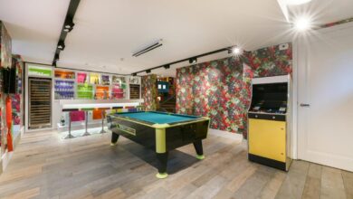 A colorful game room features a pool table, arcade cabinet, and bright bar beneath modern lighting in a basement.