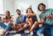 Four friends laugh and play video games together in a bright living room with snacks on the floor nearby.