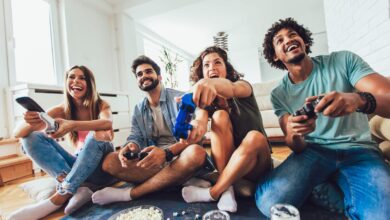 Four friends laugh and play video games together in a bright living room with snacks on the floor nearby.